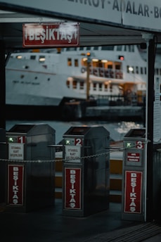Three metallic turnstiles marked with 'Beşiktaş' are positioned at a ferry terminal entrance. Behind them, a large ferry boat is illuminated, docked at the harbor. The scene is set during the evening, with dim lighting and a slightly industrial ambiance.