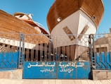 Two large wooden ships are docked behind a blue metal gate with inscriptions in Arabic. The boats are elevated, showing their full hulls, and the scene is set in an outdoor shipyard with a clear blue sky overhead.