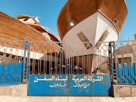 Two large wooden ships are docked behind a blue metal gate with inscriptions in Arabic. The boats are elevated, showing their full hulls, and the scene is set in an outdoor shipyard with a clear blue sky overhead.