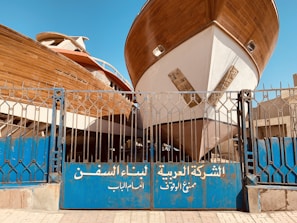 Two large wooden ships are docked behind a blue metal gate with inscriptions in Arabic. The boats are elevated, showing their full hulls, and the scene is set in an outdoor shipyard with a clear blue sky overhead.