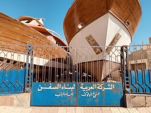 Two large wooden ships are docked behind a blue metal gate with inscriptions in Arabic. The boats are elevated, showing their full hulls, and the scene is set in an outdoor shipyard with a clear blue sky overhead.