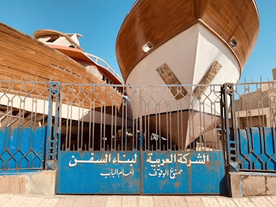 Two large wooden ships are docked behind a blue metal gate with inscriptions in Arabic. The boats are elevated, showing their full hulls, and the scene is set in an outdoor shipyard with a clear blue sky overhead.