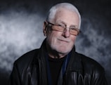An older adult with white hair, wearing glasses and a black leather jacket, is smiling gently against a textured, dark background.