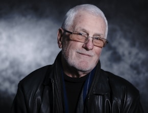 An older adult with white hair, wearing glasses and a black leather jacket, is smiling gently against a textured, dark background.