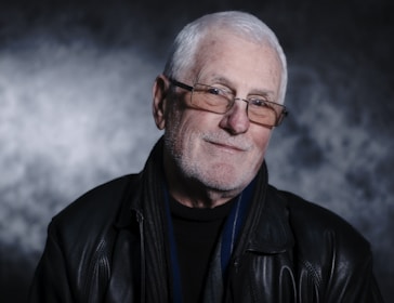 An older adult with white hair, wearing glasses and a black leather jacket, is smiling gently against a textured, dark background.