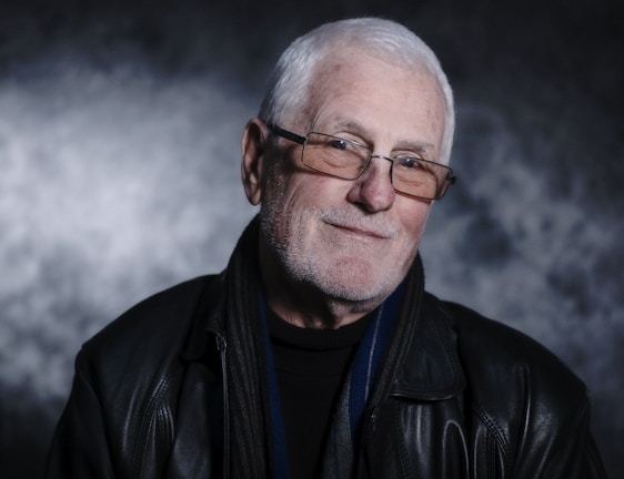 An older adult with white hair, wearing glasses and a black leather jacket, is smiling gently against a textured, dark background.