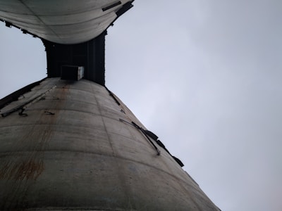 Two towering concrete silos are viewed from below, converging towards the cloudy sky. The surfaces are weathered, with visible rust stains and structural elements attached.