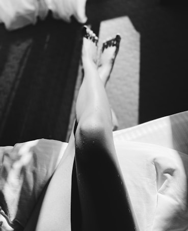 A high-contrast black and white image showing a pair of legs illuminated by sunlight. The legs have droplets of water on them, and they're partly in shadow, with light striations on the fabric they're resting on.