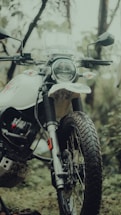 A close-up of a motorcycle in an outdoor setting, featuring a front view with visible headlight and handlebars. The motorcycle has a rugged design with knobby tires, suited for off-road travel. The background includes greenery and blurry foliage, creating a natural and adventurous atmosphere.