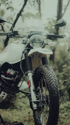 A close-up of a motorcycle in an outdoor setting, featuring a front view with visible headlight and handlebars. The motorcycle has a rugged design with knobby tires, suited for off-road travel. The background includes greenery and blurry foliage, creating a natural and adventurous atmosphere.