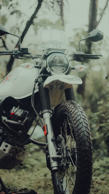 A close-up of a motorcycle in an outdoor setting, featuring a front view with visible headlight and handlebars. The motorcycle has a rugged design with knobby tires, suited for off-road travel. The background includes greenery and blurry foliage, creating a natural and adventurous atmosphere.