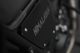 Close-up image of a motorcycle part with the word 'HIMALAYAN' printed on a black metal panel. The focus is on the branding with a blurred background that includes metallic and mechanical components.