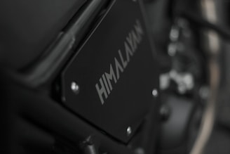 Close-up image of a motorcycle part with the word 'HIMALAYAN' printed on a black metal panel. The focus is on the branding with a blurred background that includes metallic and mechanical components.