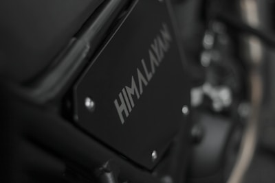 Close-up image of a motorcycle part with the word 'HIMALAYAN' printed on a black metal panel. The focus is on the branding with a blurred background that includes metallic and mechanical components.