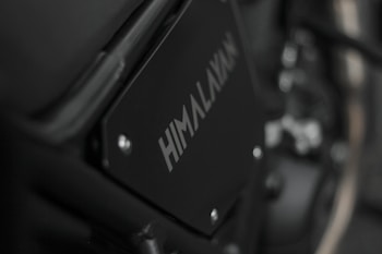 Close-up image of a motorcycle part with the word 'HIMALAYAN' printed on a black metal panel. The focus is on the branding with a blurred background that includes metallic and mechanical components.