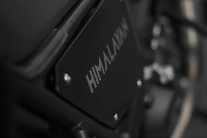 Close-up image of a motorcycle part with the word 'HIMALAYAN' printed on a black metal panel. The focus is on the branding with a blurred background that includes metallic and mechanical components.