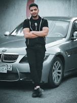 A man stands confidently with crossed arms in front of a silver BMW car. He is wearing black clothing including a jacket and pants. The car is parked on an urban street near a concrete wall, suggesting an outdoor setting.