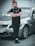 A man stands confidently with crossed arms in front of a silver BMW car. He is wearing black clothing including a jacket and pants. The car is parked on an urban street near a concrete wall, suggesting an outdoor setting.
