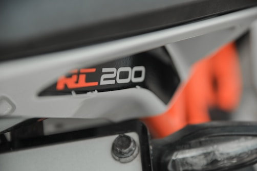 A close-up view of a motorcycle part featuring the model name RC 200 in bold white and orange text on a black background. The surrounding area appears to be part of the motorcycle's frame and components, with visible screws and a portion of orange piping or framework.