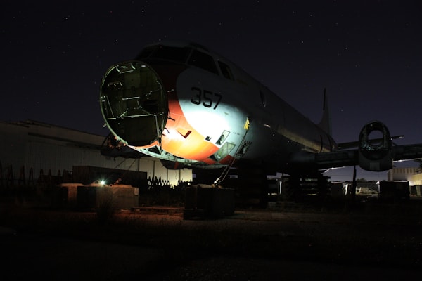 A partially disassembled airplane with the nose cone removed is situated in a dark, possibly abandoned setting. It is propped up on blocks, and some parts are missing, exposing fixtures and structures. The atmosphere is dimly lit, with faint illuminate on the body's side revealing the number 357.