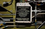 A warning instruction plate with text indicating filling instructions for an emergency brake reservoir. It lists pressure specifications, temperature rise warnings, and a minimum charge requirement. Surrounding the plate are various metal pipes, bolts, and components of a mechanical system.