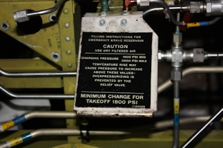 A warning instruction plate with text indicating filling instructions for an emergency brake reservoir. It lists pressure specifications, temperature rise warnings, and a minimum charge requirement. Surrounding the plate are various metal pipes, bolts, and components of a mechanical system.