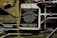 A warning instruction plate with text indicating filling instructions for an emergency brake reservoir. It lists pressure specifications, temperature rise warnings, and a minimum charge requirement. Surrounding the plate are various metal pipes, bolts, and components of a mechanical system.