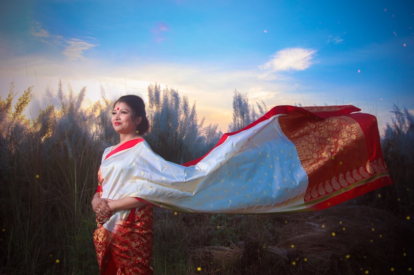 A woman wearing a traditional red and white sari stands gracefully in a field of tall grasses under a vibrant sky. Her attire features intricate gold designs, and she seems to be enjoying a serene moment as the fabric of her sari catches the wind. The background is filled with fluffy reeds and a colorful sunset, enhancing the peaceful ambiance.