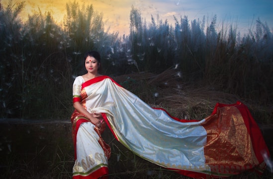 A woman dressed in a traditional white and red saree sits gracefully amidst tall grass. The saree features gold detailing, and she wears a bindi on her forehead. The background shows a warm sunset or sunrise, providing a serene and natural atmosphere.