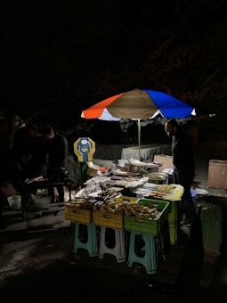 A nighttime market scene with a small food stall. Skewers are displayed on several colorful crates and stools, all set under a large umbrella with red, blue, and white panels. Two people are seen near the stall, one of them is preparing food on a grill. The surrounding area is dimly lit, creating a cozy street food ambiance.