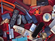 A variety of disparate objects are arranged on a red fabric surface, including multiple old mobile phones, a television remote control, a fidget spinner, a razor in an orange case, and several leather wallets. Other items include a shoe brush, writing pens, a small MP3 player, and a bar of soap. The collection features miscellaneous everyday items and novelties.