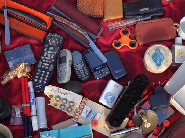 A variety of disparate objects are arranged on a red fabric surface, including multiple old mobile phones, a television remote control, a fidget spinner, a razor in an orange case, and several leather wallets. Other items include a shoe brush, writing pens, a small MP3 player, and a bar of soap. The collection features miscellaneous everyday items and novelties.