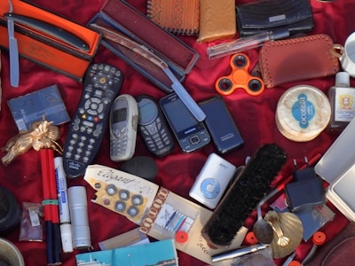A variety of disparate objects are arranged on a red fabric surface, including multiple old mobile phones, a television remote control, a fidget spinner, a razor in an orange case, and several leather wallets. Other items include a shoe brush, writing pens, a small MP3 player, and a bar of soap. The collection features miscellaneous everyday items and novelties.
