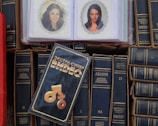 A collection of books with dark covers and Cyrillic writing is displayed with a focus on the works of Lenin. In the center lies a videocassette cover with distinctive typography and graphic symbols. Two laminated photographs of women are visible on top of the books.