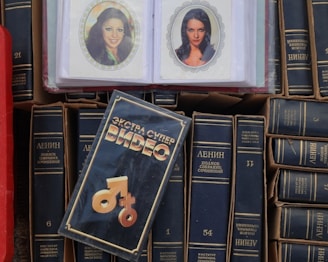 A collection of books with dark covers and Cyrillic writing is displayed with a focus on the works of Lenin. In the center lies a videocassette cover with distinctive typography and graphic symbols. Two laminated photographs of women are visible on top of the books.