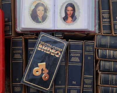 A collection of books with dark covers and Cyrillic writing is displayed with a focus on the works of Lenin. In the center lies a videocassette cover with distinctive typography and graphic symbols. Two laminated photographs of women are visible on top of the books.