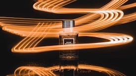 A bottle of perfume is placed against a dark background. Surrounding it are dynamic, swirling light trails in shades of orange and yellow that create a sense of motion and energy.