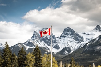 A Canadian flag is seen waving on a flagpole in the foreground with a dramatic backdrop of snow-capped mountains. Evergreen trees dot the landscape, and the scene is under a partly cloudy sky with some sunlight breaking through.