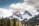 A Canadian flag is seen waving on a flagpole in the foreground with a dramatic backdrop of snow-capped mountains. Evergreen trees dot the landscape, and the scene is under a partly cloudy sky with some sunlight breaking through.