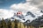 A Canadian flag is seen waving on a flagpole in the foreground with a dramatic backdrop of snow-capped mountains. Evergreen trees dot the landscape, and the scene is under a partly cloudy sky with some sunlight breaking through.