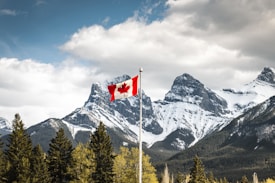 A Canadian flag is seen waving on a flagpole in the foreground with a dramatic backdrop of snow-capped mountains. Evergreen trees dot the landscape, and the scene is under a partly cloudy sky with some sunlight breaking through.
