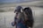 A person with long hair, wearing a blue shirt adorned with red dachshund patterns, is holding a gray Weimaraner puppy outdoors. The background features an expansive, blurred landscape with muted green and brown tones, suggesting a rural or countryside setting.