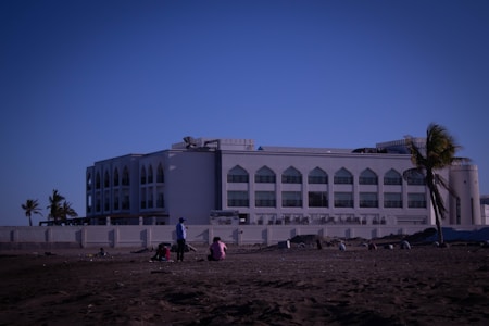 A large white building with arched windows, likely institutional or government-related, situated near a sandy area with several people sitting or standing. Palm trees are present, adding a tropical feel.