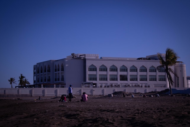 A large white building with arched windows, likely institutional or government-related, situated near a sandy area with several people sitting or standing. Palm trees are present, adding a tropical feel.
