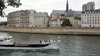 A river scene featuring a tourist boat navigating the waterway, with people on board. The background is dominated by historic buildings, showcasing classic European architecture typical of a cityscape. Prominent is a cathedral with a tall spire, contributing to the historical ambiance. Leafy trees border the riverbank, adding a touch of nature.