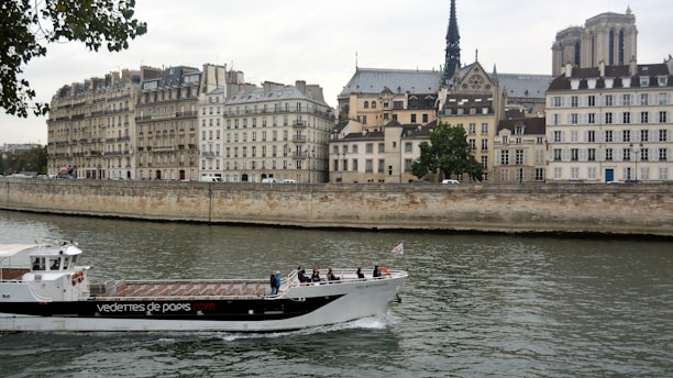 A river scene featuring a tourist boat navigating the waterway, with people on board. The background is dominated by historic buildings, showcasing classic European architecture typical of a cityscape. Prominent is a cathedral with a tall spire, contributing to the historical ambiance. Leafy trees border the riverbank, adding a touch of nature.