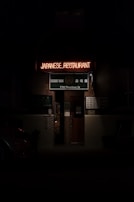 An entrance to a Japanese restaurant is seen with a glowing neon sign reading 'JAPANESE RESTAURANT.' Beneath, a green sign displays 'Sushi Yan' and the address '770 Thurlow St.' The image is dimly lit, creating a moody atmosphere.