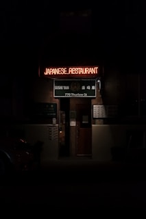 An entrance to a Japanese restaurant is seen with a glowing neon sign reading 'JAPANESE RESTAURANT.' Beneath, a green sign displays 'Sushi Yan' and the address '770 Thurlow St.' The image is dimly lit, creating a moody atmosphere.