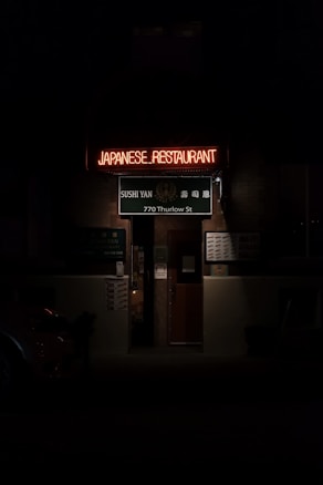 An entrance to a Japanese restaurant is seen with a glowing neon sign reading 'JAPANESE RESTAURANT.' Beneath, a green sign displays 'Sushi Yan' and the address '770 Thurlow St.' The image is dimly lit, creating a moody atmosphere.