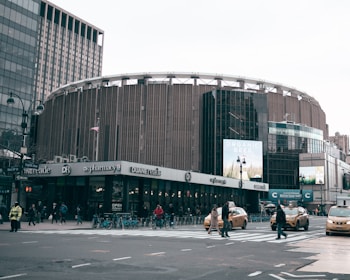 A large, round building with a modern design dominates the urban streetscape. The structure houses various commercial establishments like pharmacies, and a billboard advertising organic beer can be seen. Numerous pedestrians cross the busy intersection, while yellow taxis and bicycles add to the city's bustling atmosphere.